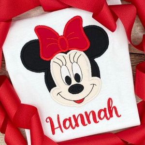 May include: White t-shirt with a red and black embroidered Minnie Mouse design and the name "Hannah" in red embroidery.