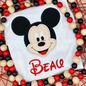 Puede incluir: Camiseta blanca con una cabeza de Mickey Mouse bordada y el nombre "Beau" en rojo.