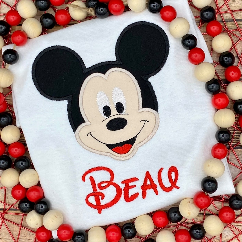 Puede incluir: Camiseta blanca con una cabeza de Mickey Mouse bordada y el nombre "Beau" en rojo.