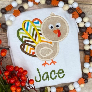 May include: White t-shirt featuring a cartoon turkey with a beige body, colorful tail feathers, and the name "Jace" in green script. The turkey has a red wattle and orange feet. The shirt is decorated with a Thanksgiving theme.