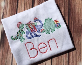 May include: White t-shirt with an embroidered design of three dinosaurs wearing Christmas hats and holding presents. The name "Ben" is embroidered below the dinosaurs.