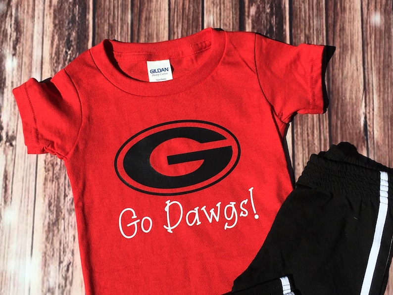 Puede incluir: Camiseta roja de manga corta con un gr&aacute;fico en blanco y negro de una 'G' dentro de un &oacute;valo y el texto 'Go Dawgs!'