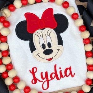 May include: White t-shirt with an embroidered image of Minnie Mouse with a red bow and the name Lydia in red lettering.