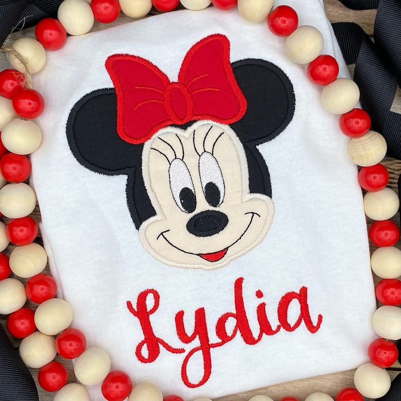 May include: White t-shirt with an embroidered image of Minnie Mouse with a red bow and the name Lydia in red lettering.