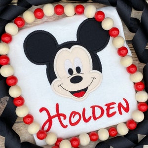 Puede incluir: Camiseta blanca con una imagen bordada de Mickey Mouse y el nombre "Holden" en rojo.