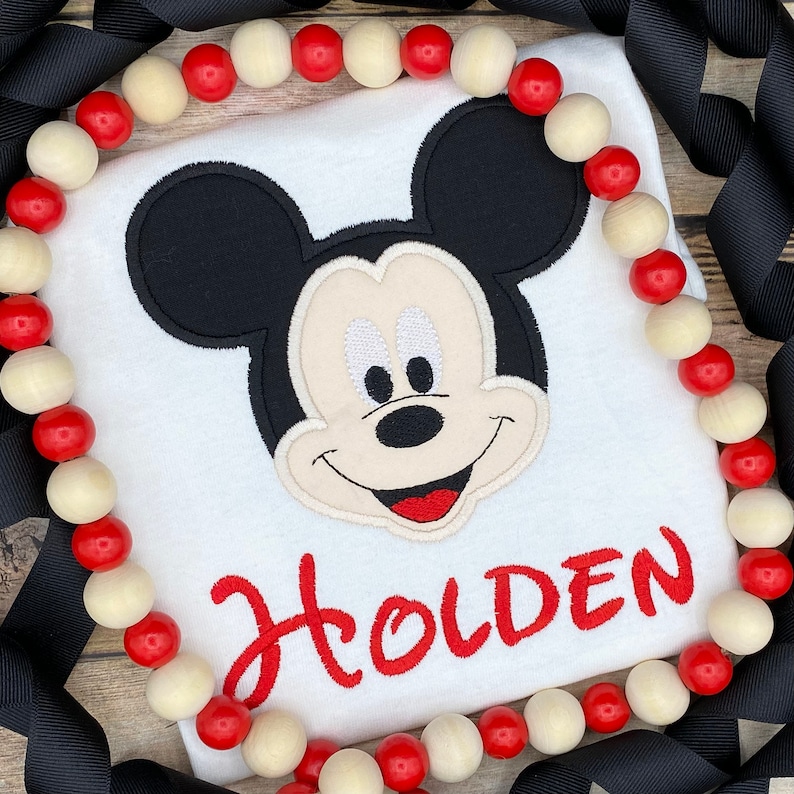 Puede incluir: Camiseta blanca con una imagen bordada de Mickey Mouse y el nombre "Holden" en rojo.