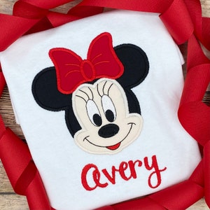 May include: White t-shirt with a red and black embroidered Minnie Mouse design and the name "Avery" in red embroidery.