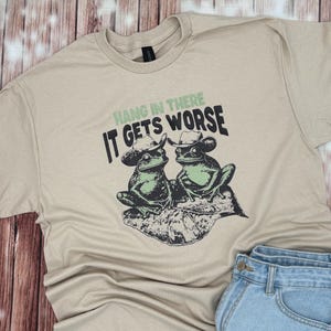 May include: Beige t-shirt featuring a graphic of two frogs wearing cowboy hats, sitting on a leaf. The text "Hang in there, it gets worse" is printed above the frogs in green and black.