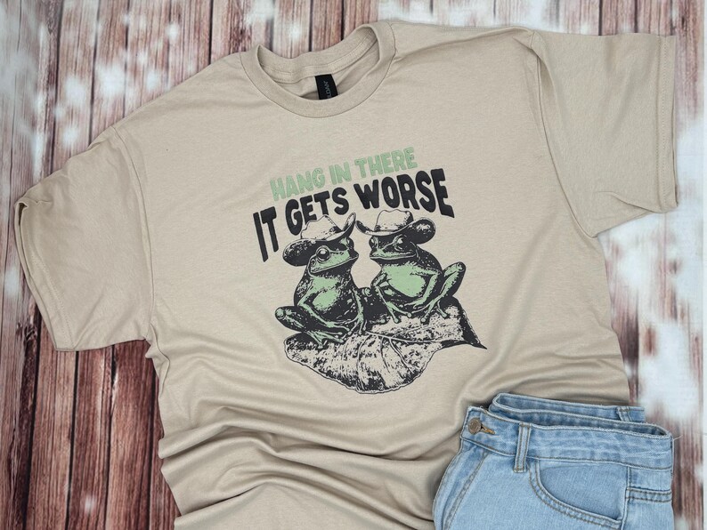 May include: Beige t-shirt featuring a graphic of two frogs wearing cowboy hats, sitting on a leaf. The text "Hang in there, it gets worse" is printed above the frogs in green and black.