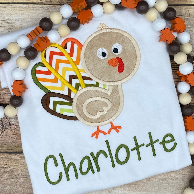 May include: White t-shirt with an embroidered turkey design. The turkey has a tan body, a red wattle, and a colorful tail. The name "Charlotte" is embroidered in green below the turkey. Decorative wooden beads surround the shirt.