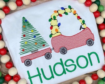 May include: White Christmas-themed t-shirt with a red wagon pulling a green Christmas tree. The wagon is decorated with colorful lights and the name "Hudson" is embroidered in green below the wagon.