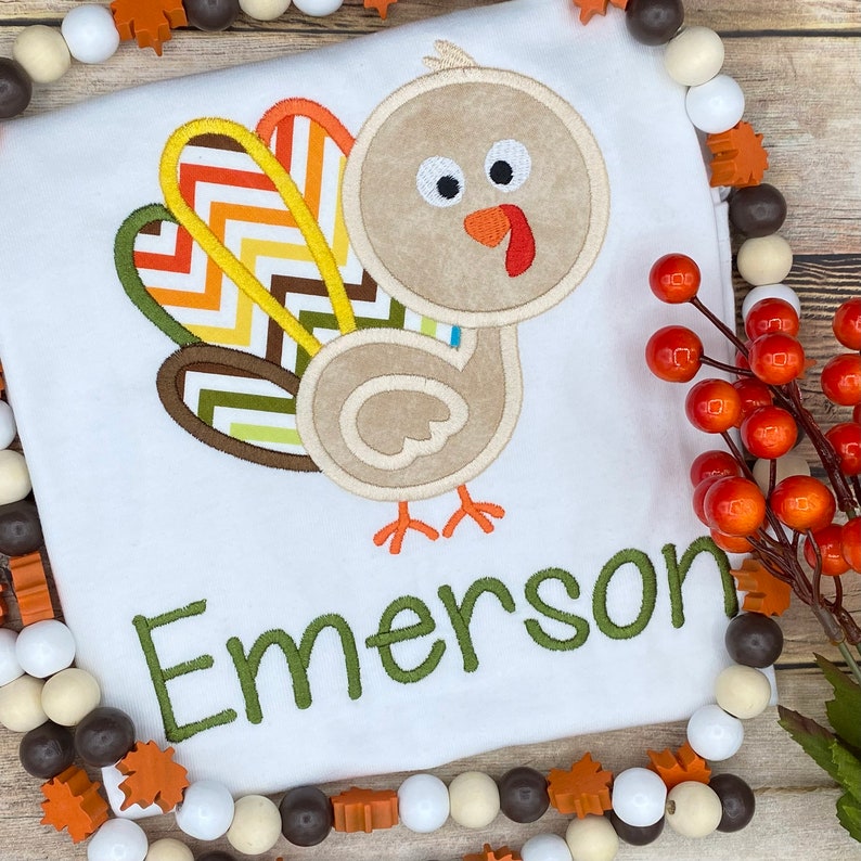 May include: White t-shirt featuring a cartoon turkey applique with colorful feathers and the name "Emerson" embroidered in green. The turkey has a tan body, orange beak and feet, and black eyes. Thanksgiving themed apparel.