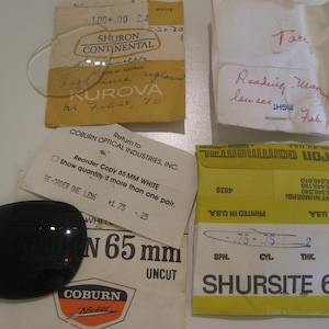 May include: An assortment of vintage eyeglass lenses and packaging. Includes a clear lens, a dark tinted lens, and several paper envelopes and cards with handwritten notes and printed text. The packaging features the brands "Shuron Continental" and "Coburn Optical Industries."