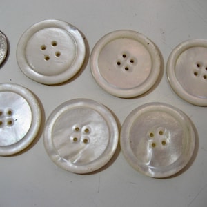 May include: A collection of seven off-white, round buttons with four holes each. The buttons vary slightly in size and have a pearlescent sheen. A quarter is included for size comparison.