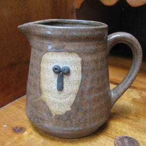 May include: A brown ceramic pitcher with a white face and black eyes. The pitcher has a rustic, handmade look.