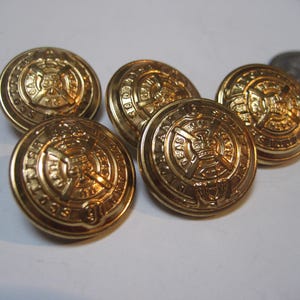 May include: A collection of six gold-colored metal buttons. Each button has a detailed embossed design featuring a central emblem surrounded by text. The buttons are round and have a shiny finish, suggesting they are new or well-preserved.