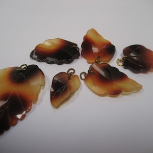 May include: Six leaf-shaped pendants with a gradient of colors from light yellow to dark brown. Each leaf has a gold-colored loop at the top for hanging. The leaves vary in size and shape, with detailed veining.