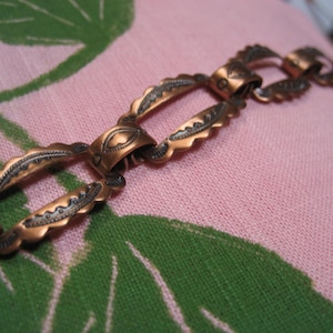 May include: Close-up of a copper-colored chain with ornate, scalloped links. The chain is resting on a pink fabric with a large green leaf pattern. The links have a detailed, textured design.