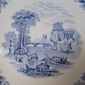 May include: Blue and white transferware plate with a scenic design of a building, bridge, and trees. The design is in a blue and white color scheme.