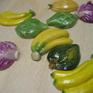 May include: A collection of ceramic vegetable figurines, including bananas, artichokes, and cabbages. The bananas are yellow, the artichokes are green, and the cabbages are purple. The figurines are arranged on a light-colored wooden surface.