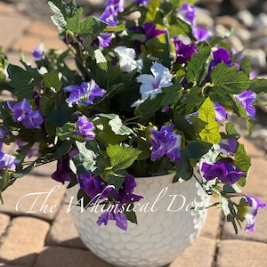 May include: A white textured ceramic pot filled with vibrant purple and white flowers and green foliage. The pot has a unique, dimpled surface. The text "The Whimsical Door" is written on the pot.