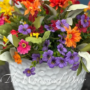 May include: A white ceramic planter with a textured surface, filled with a colorful arrangement of artificial flowers. The flowers include orange, red, yellow, and purple blooms. The text "The Whimsical Door" is printed on the planter.