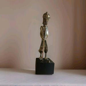 May include: A brass sculpture of a golfer in a stooped stance, holding a golf club. The figure is mounted on a black square base. The sculpture has a unique, stylized design.