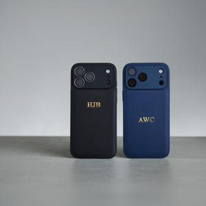 May include: Two textured phone cases, one black and one blue, each featuring a camera cutout and gold embossed initials. The black case displays the letters HJB, while the blue case displays the letters AWC. Both cases have a leather-like finish.