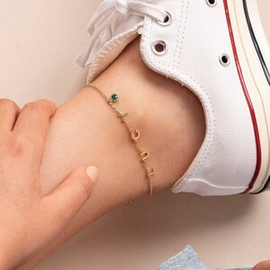 Peut inclure: Un bracelet de cheville en or avec un petit charme en pierre verte et des charmes en lettres. Le bracelet est porté à la cheville d'une personne avec des baskets blanches et un jean bleu.