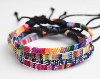 May include: Three colorful woven bracelets with black adjustable cords. The bracelets are layered on top of each other and feature a variety of colors, including red, orange, yellow, green, blue, purple, and pink.