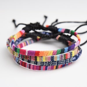 May include: Three colorful woven bracelets with black adjustable cords. The bracelets are layered on top of each other and feature a variety of colors, including red, orange, yellow, green, blue, purple, and pink.