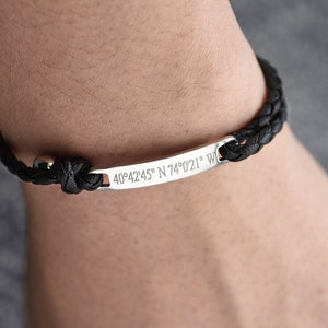 May include: A black leather braided bracelet with a silver bar engraved with the coordinates "40°42'45" N 74°0'21" W".