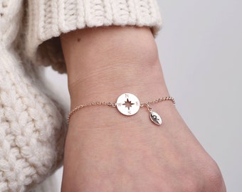 May include: A delicate gold chain bracelet with a compass charm and a small leaf charm. The compass charm has a star in the center and the letters N, S, E, and W around the edge.