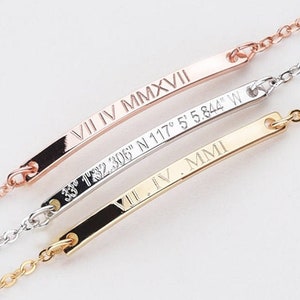 May include: Three delicate gold, silver, and rose gold bar bracelets with engraved Roman numerals and coordinates. The bracelets are layered on a white background.