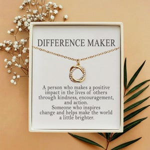 Könnte beinhalten: Eine goldfarbene Halskette mit einem kreisförmigen Anhänger, der mit kleinen klaren Steinen besetzt ist, auf einer weißen Karte mit dem Text "DIFFERENCE MAKER" und einer Definition, präsentiert in einer quadratischen Schachtel.