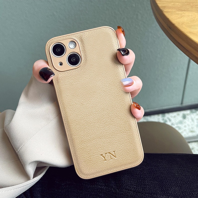 May include: A beige smartphone case with a textured, pebbled surface finish. The case features a square camera cutout with three circular lenses and a flash. The letters YN are embossed in a serif font at the bottom centre of the back panel.