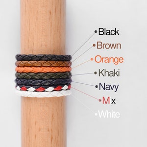 May include: A stack of braided leather bracelets in various colors, including black, brown, orange, khaki, navy, white, and a mix of colors. The bracelets are displayed on a wooden dowel.