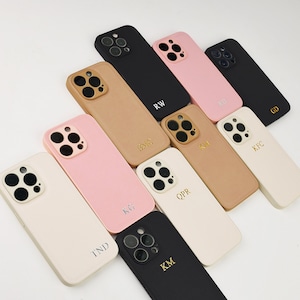 May include: A collection of ten smartphone cases in black, tan, pink, and cream colours. Each case features a triple-lens camera cutout and is personalised with two or three gold or silver embossed letters, including RW, RD, GD, BMC, KM, KFC, KG, QPR, TND, and KM.