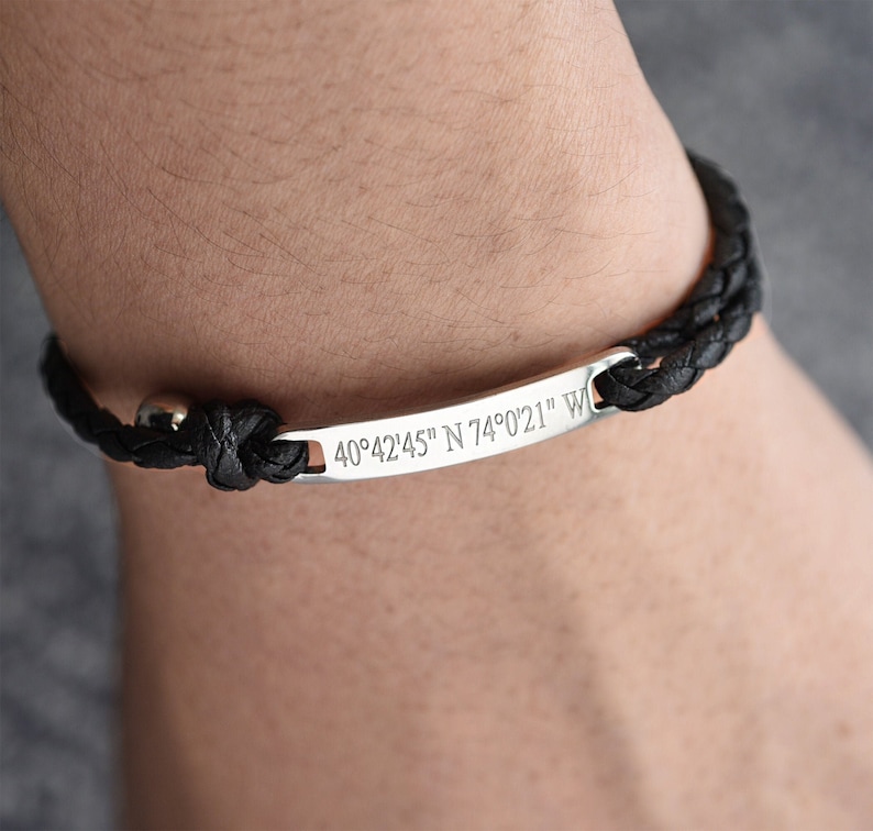 May include: A black leather braided bracelet with a silver bar engraved with the coordinates "40&deg;42'45" N 74&deg;0'21" W".