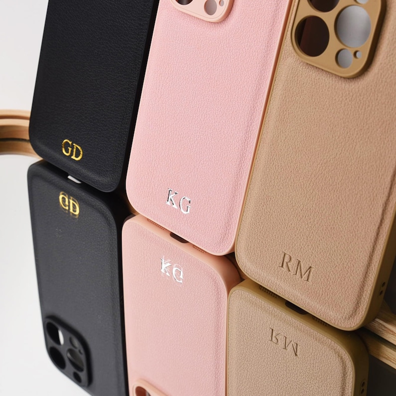 May include: A collection of textured smartphone cases in black, light pink, and tan. Each case features a camera cutout and a two-letter monogram embossed in gold or silver foil. The cases are arranged in a grid, with reflections visible below.