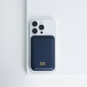 May include: A navy blue leather phone wallet with the initials "BK" embossed in gold. The wallet is attached to the back of a white iPhone.