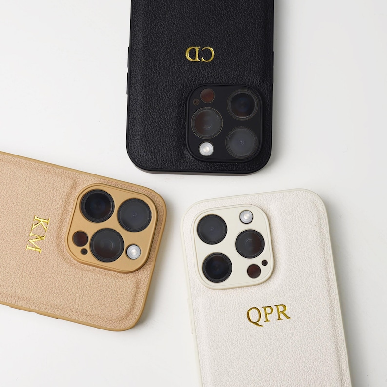 May include: Three textured leather phone cases in black, tan, and cream. Each case features a triple-lens camera cutout and gold foil-stamped initials: CD on the black case, KM on the tan case, and QPR on the cream case. The cases have a pebbled finish.