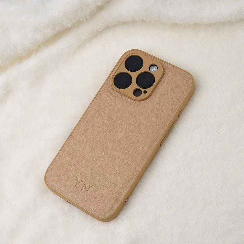 May include: A tan-coloured smartphone case with a textured surface and a raised camera module featuring three circular lenses and a flash. The letters YN are embossed in the bottom left corner of the case, which has a smooth, uniform finish.