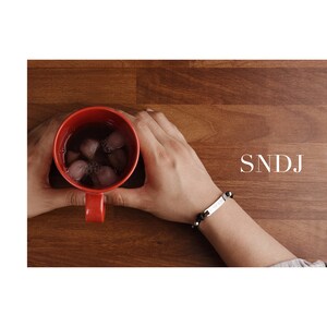 May include: A person wearing a black beaded bracelet with a silver nameplate engraved with the letters "SNDJ". The person is holding a red mug filled with ice and a dark liquid.