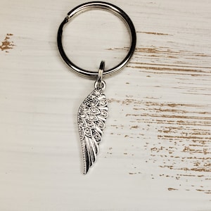 May include: A silver-colored keychain with a decorative wing charm. The wing charm is detailed with small, clear crystals and is attached to a large, round key ring. The background is a white, distressed wood surface.