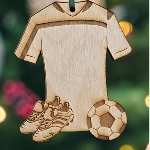 May include: Wooden soccer-themed ornament featuring a soccer jersey, cleats, and a soccer ball. The ornament is made of light-colored wood with etched details and a green ribbon for hanging. A festive decoration for soccer fans.