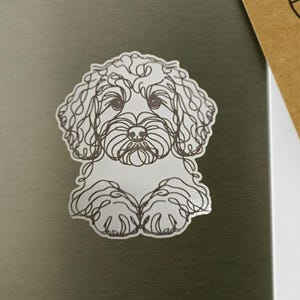 May include: A white sticker featuring a line drawing of a dog, with brown outlines. The dog has curly fur and is sitting down. The sticker is on a gray surface.