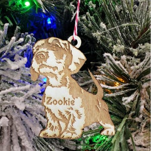 May include: A wooden Christmas ornament shaped like a dog with the name "Zookie" engraved on it. The ornament is light brown and has a white string attached to it.