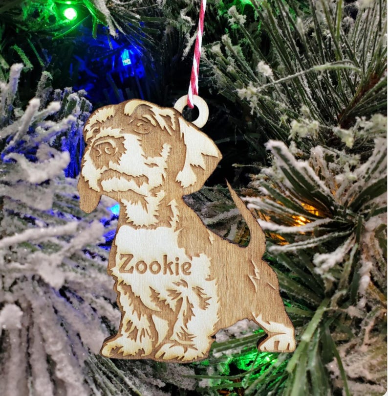 May include: A wooden Christmas ornament shaped like a dog with the name "Zookie" engraved on it. The ornament is light brown and has a white string attached to it.