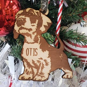 May include: A wooden Christmas ornament shaped like a dachshund dog with the name "Otis" engraved on it. The ornament is hanging on a Christmas tree with red and white candy canes and other ornaments.